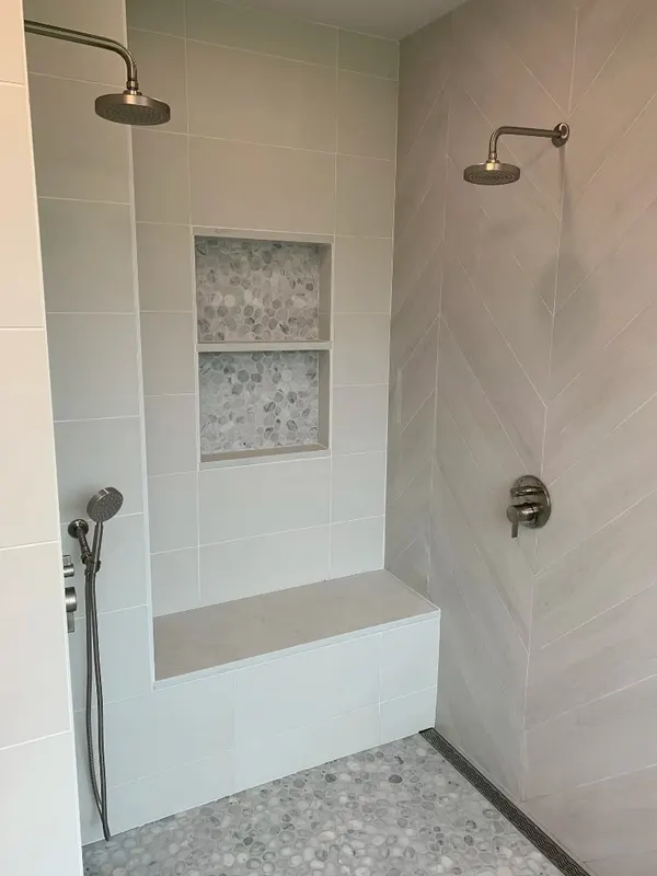 Luxury Walk-in Shower — Chevron stone walls, pebble mosaic floor, double niche, and a built-in bench with linear drain.