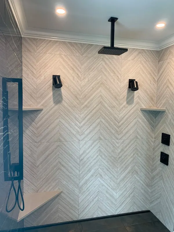 Chevron Stone Shower — Vertical chevron stone tile with matte black rainfall showerheads and integrated corner shelves.