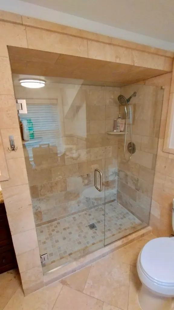 Travertine Shower with Glass Enclosure — Full-height travertine walls with a mosaic shower floor, corner shelf, and frameless glass enclosure.
