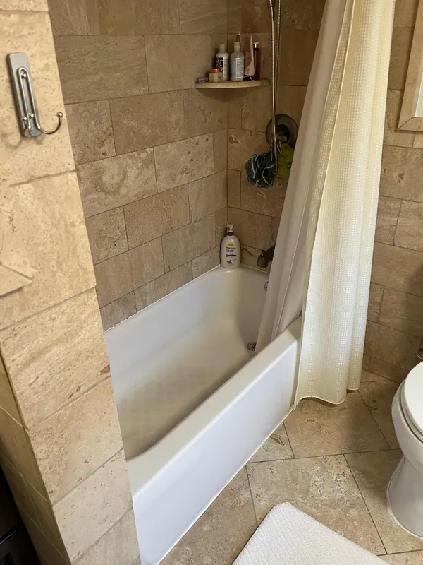 Travertine Tub & Shower Combination — Travertine walls and floor unified across the tub alcove, finished with a stone corner shelf.